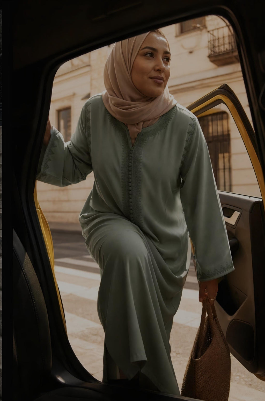 A woman wearing a sage green two-piece abaya set and a beige hijab steps out of a yellow car, holding a brown woven handbag.